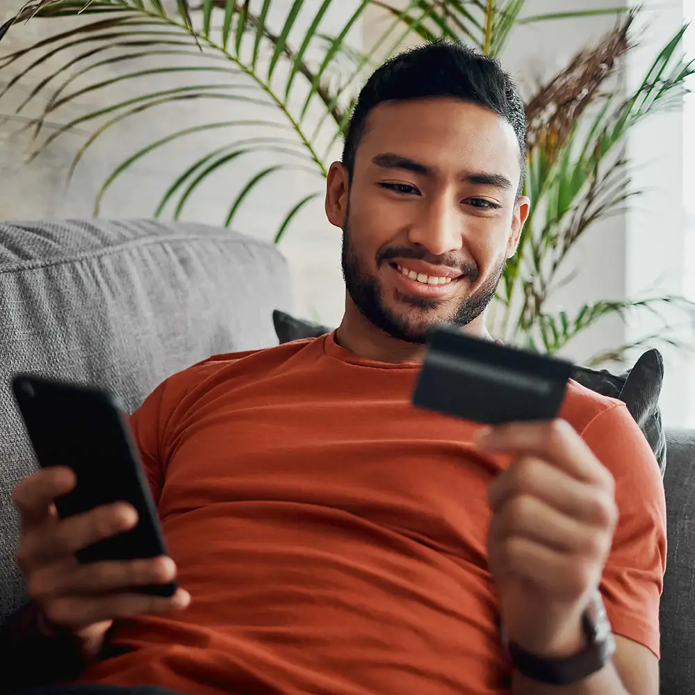 Man sitting on his couch smiling while holding his phone and looking at his debit card.